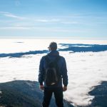 A man with a backpack stands on a mountain top, overlooking a valley covered in clouds under a bright blue sky.