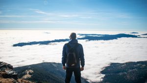 A man with a backpack stands on a mountain top, overlooking a valley covered in clouds under a bright blue sky.